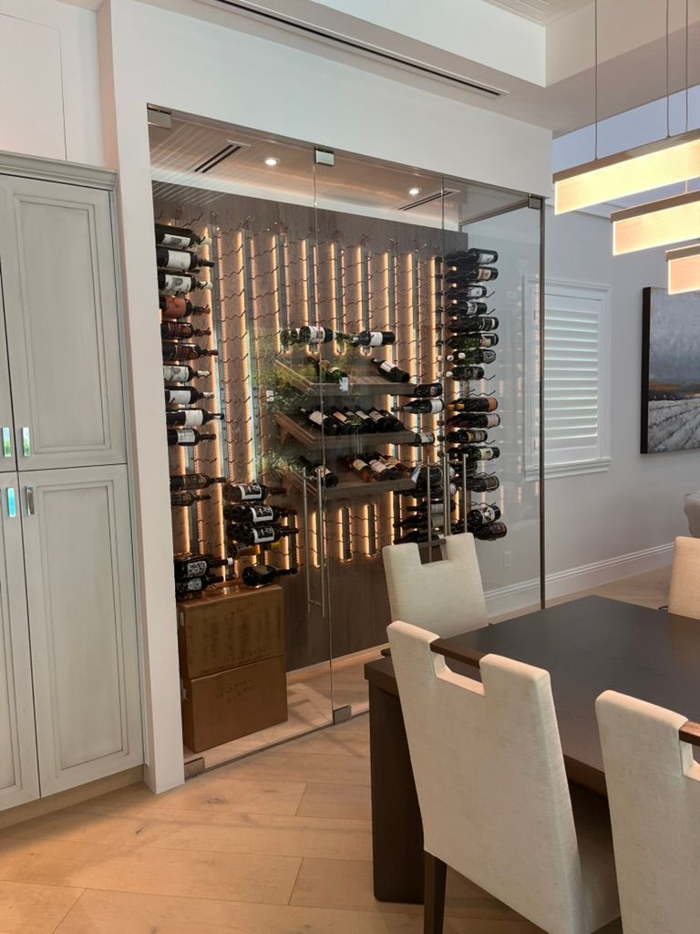 A contemporary glass-enclosed wine cellar with illuminated shelving next to a dining area with chairs and a table.