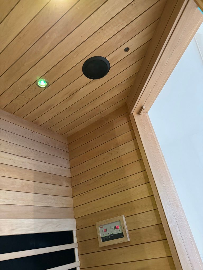 An interior view of a sauna with wooden walls, a ceiling-mounted speaker, a small green light, and a control panel.