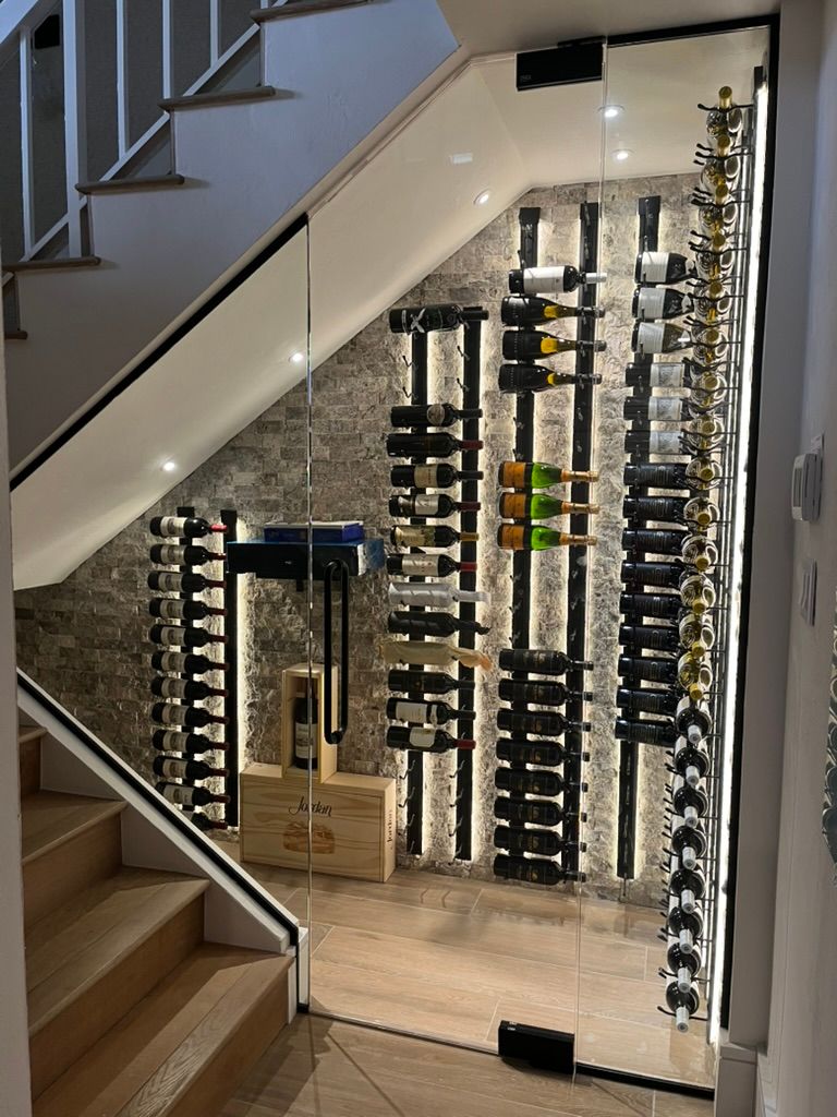 A modern glass-enclosed wine cellar built into the space underneath a staircase, featuring illuminated wall-mounted racks.