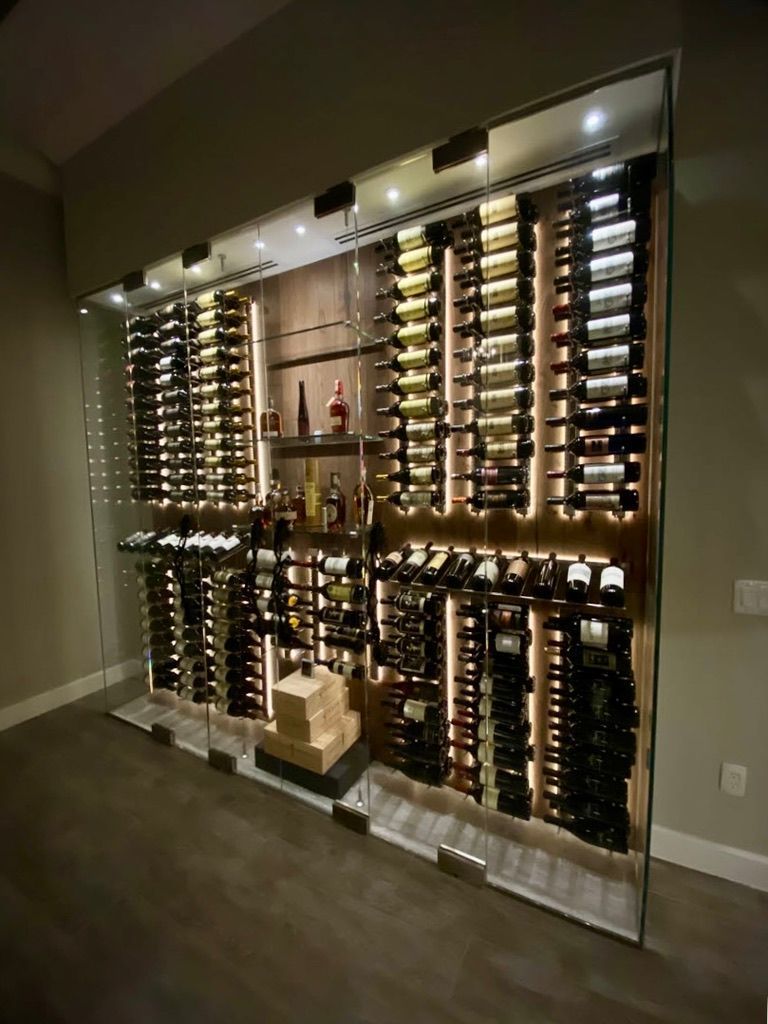 A modern, glass-enclosed wine cellar with illuminated vertical racks holding numerous bottles against a wooden back wall.