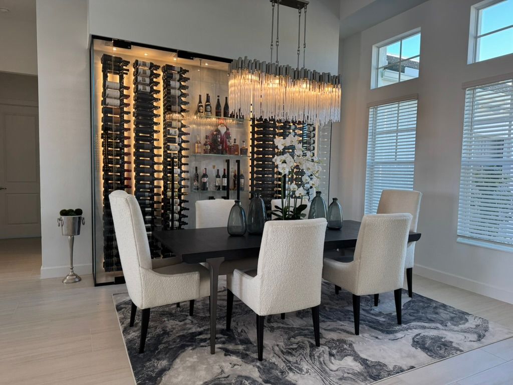 A dining room with a long table, four upholstered chairs, a crystal chandelier, and a built-in illuminated wine rack wall.