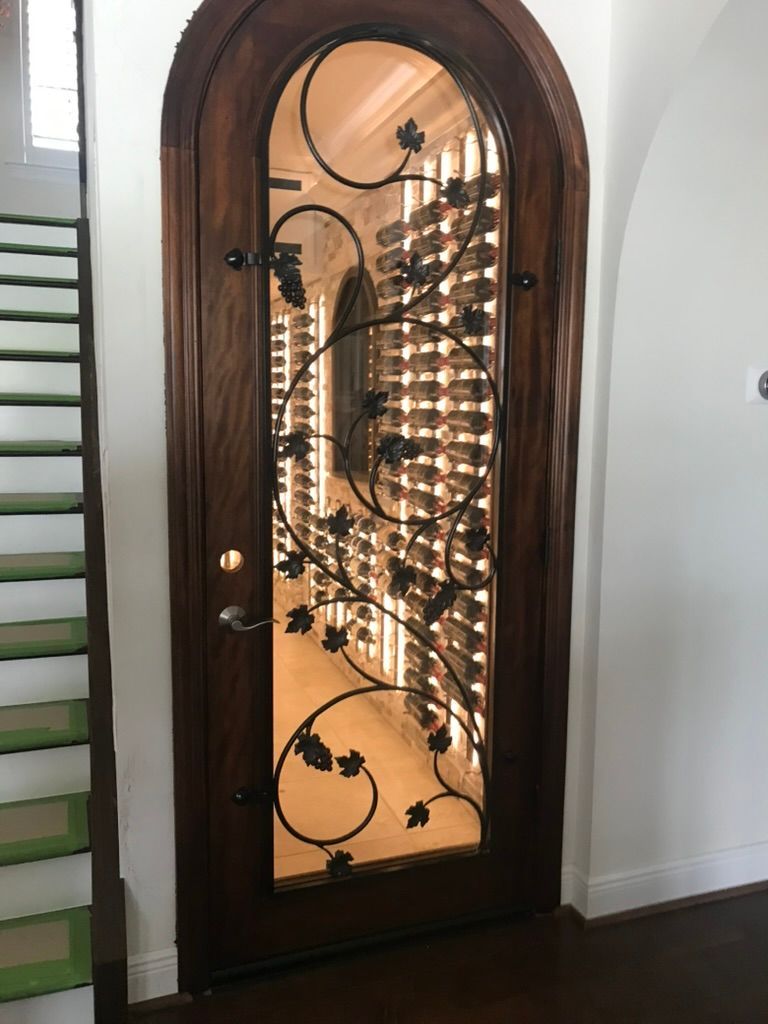 An arched wood door with glass and a decorative black metal vine grill, revealing a well-lit wine cellar inside.