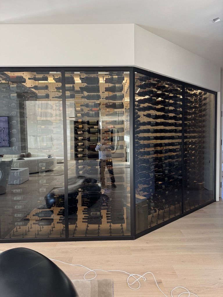A glass-walled walk-in wine cellar with dark metal shelving holding numerous wine bottles in a bright, modern room.
