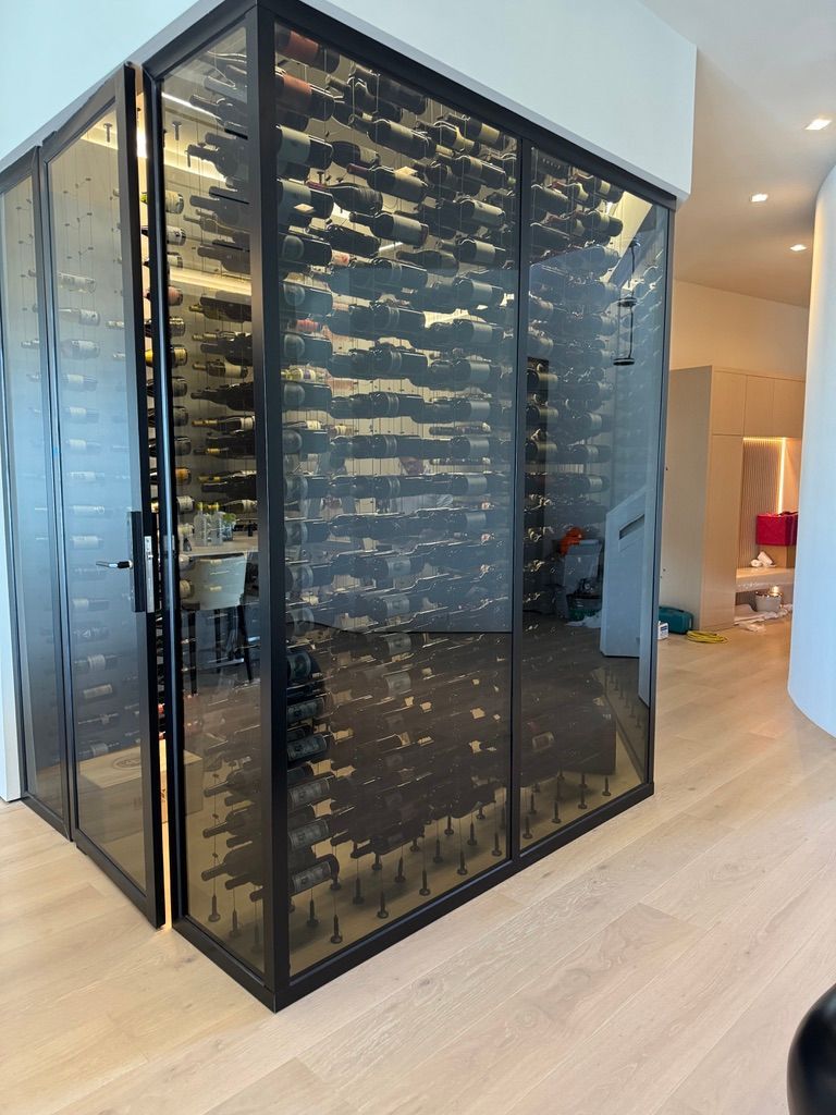 A sleek, glass-enclosed wine cellar with black metal framing stands in a modern room with light wood flooring.