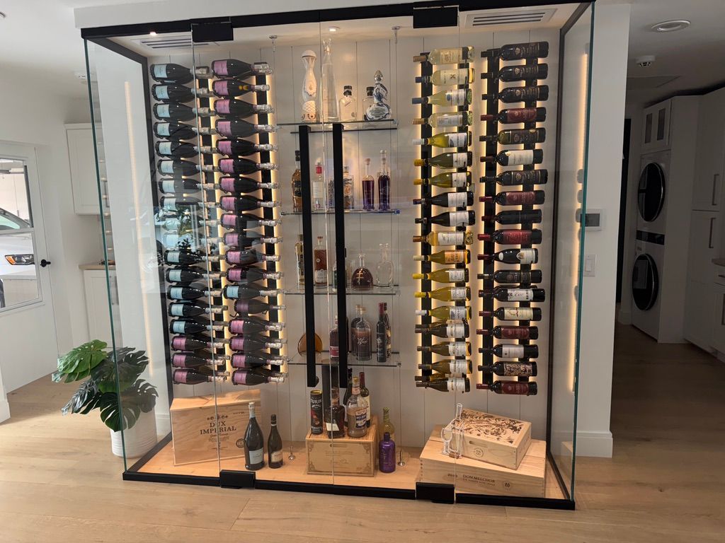 A large, modern glass wine cellar display featuring organized wine racks, illuminated shelving, and wooden storage crates.