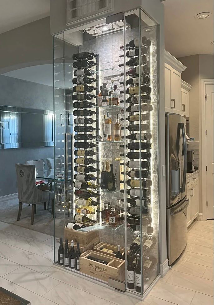 A floor-to-ceiling, glass-enclosed wine display tower integrated into a kitchen corner, filled with bottles on metal racks.