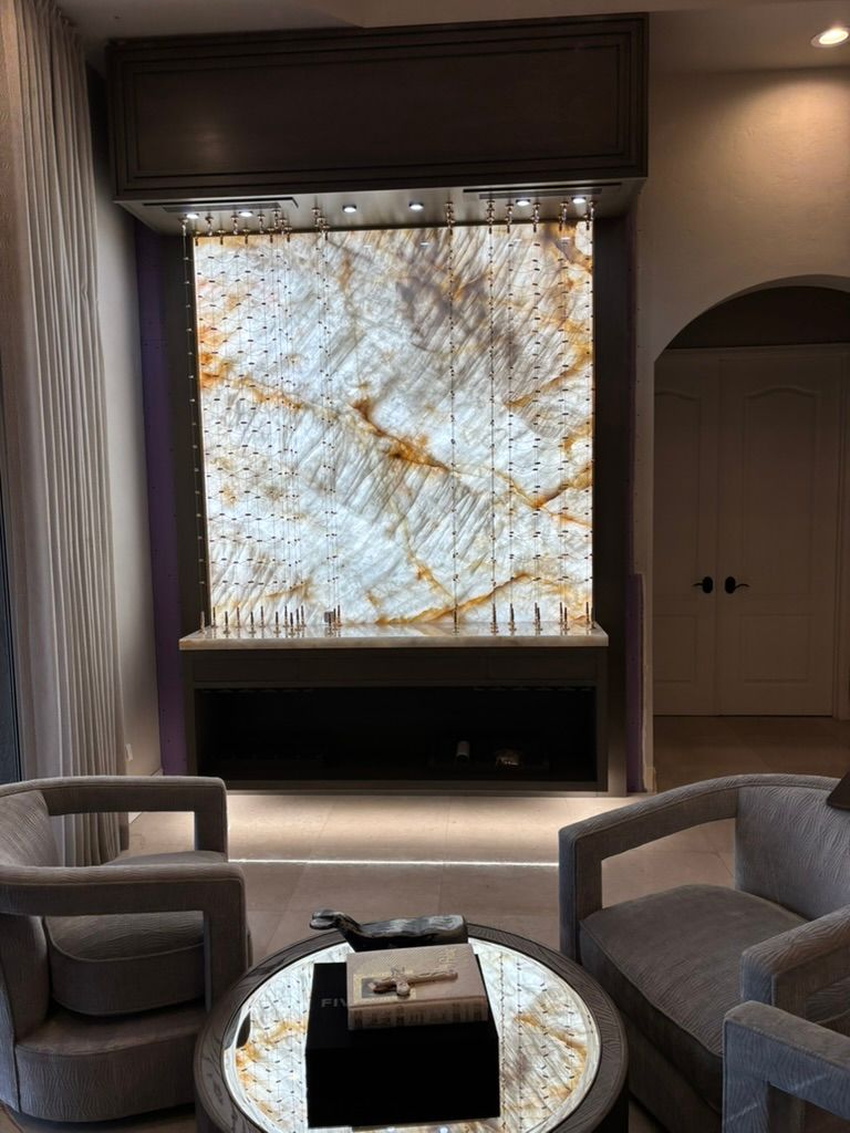 A room with two grey armchairs, a round marble coffee table, and an illuminated, marbled stone feature wall behind a bar.