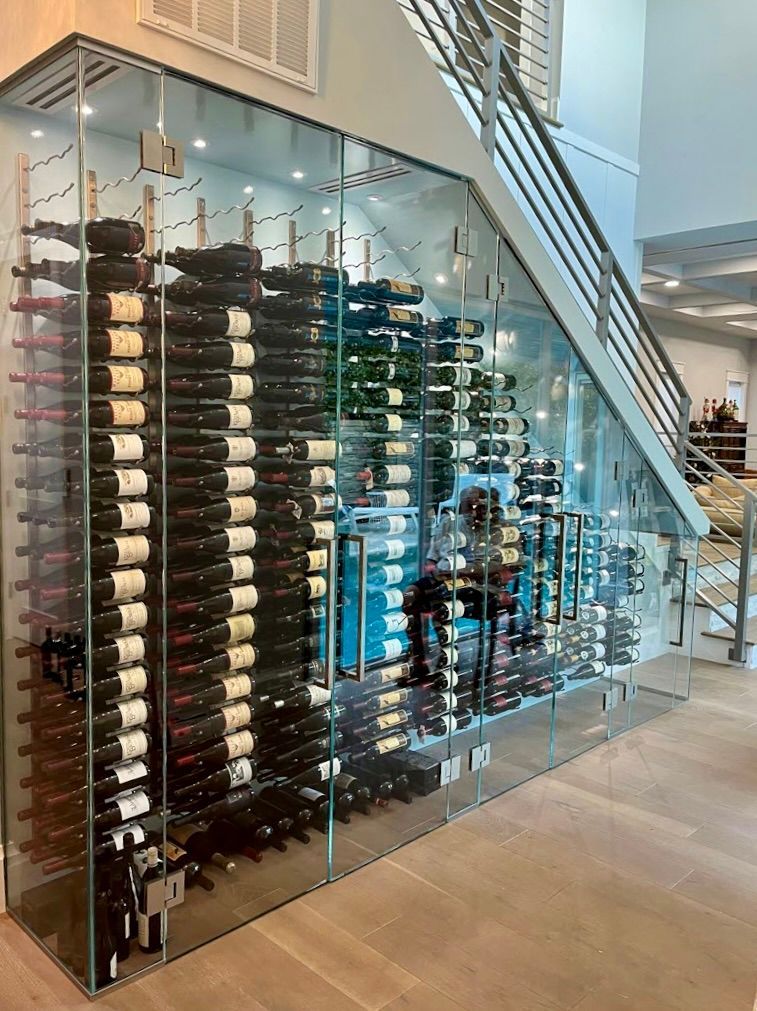 A modern, glass-enclosed wine cellar built under a staircase, featuring rows of wine bottles stored on metal racks.