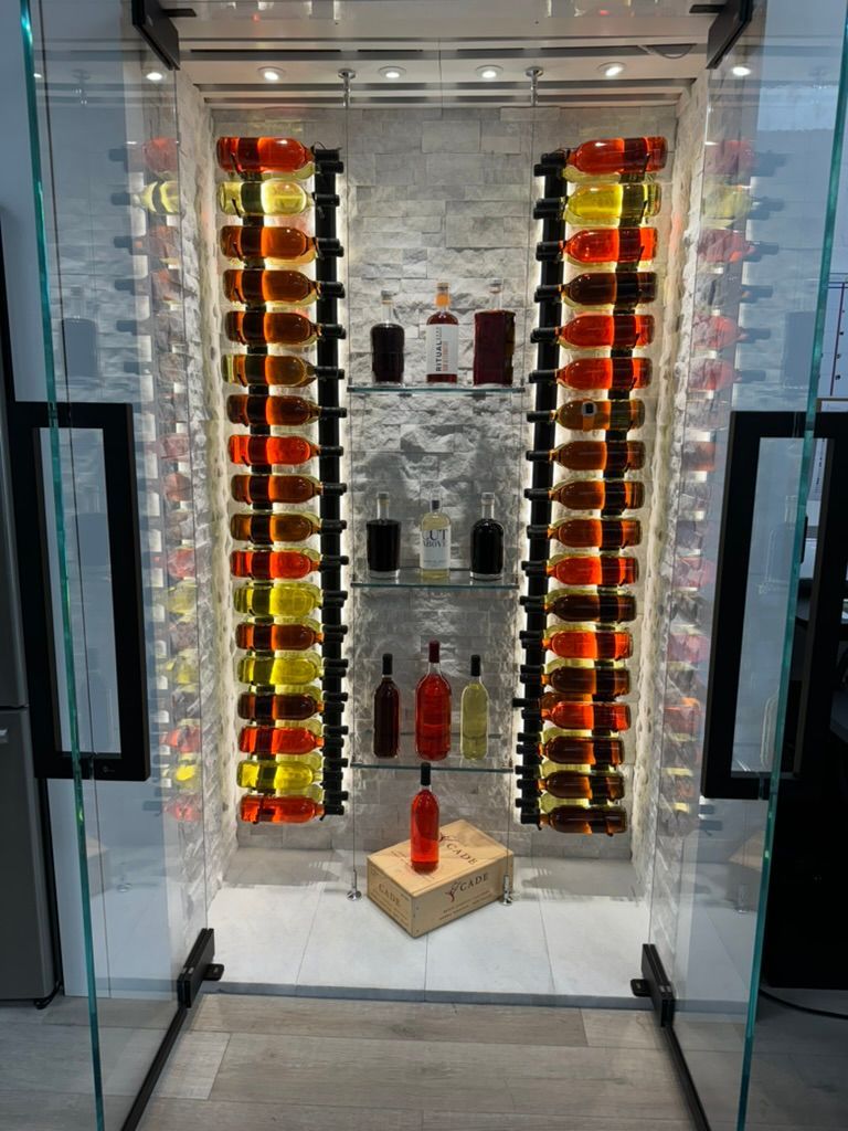 A modern glass-enclosed wine cellar with illuminated vertical bottle racks flanking a stone wall and display shelves.