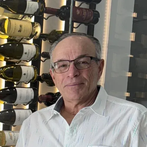 A person wearing glasses and a white button-down shirt standing in front of a wine rack.