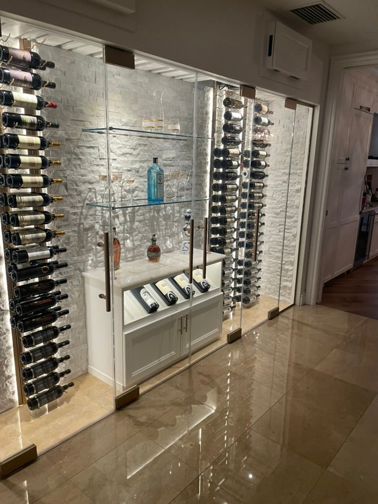 A wall-mounted wine cellar with glass doors, illuminated shelving, and rows of wine bottles on a textured white wall.
