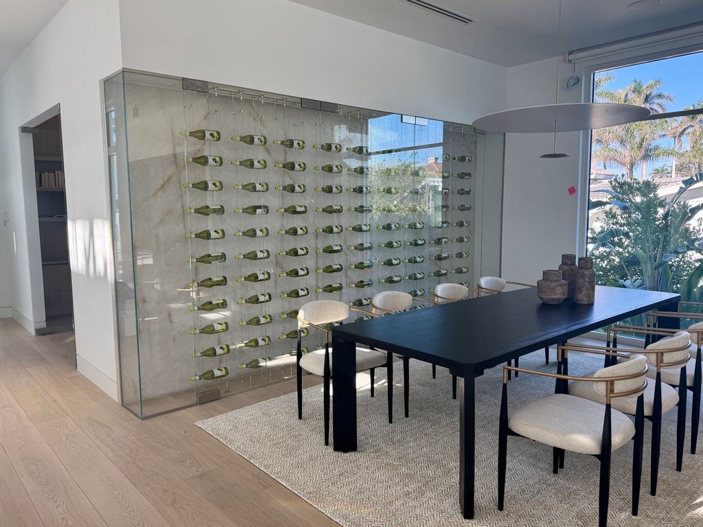 A glass-enclosed wine rack displays bottles on a light-colored wall beside a modern dining table with six chairs.