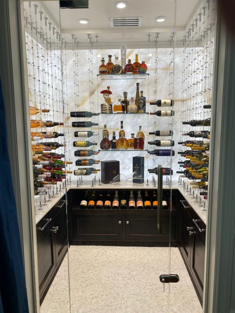 A modern wine cellar behind a glass door, featuring backlit shelves, hanging racks, and dark cabinets.