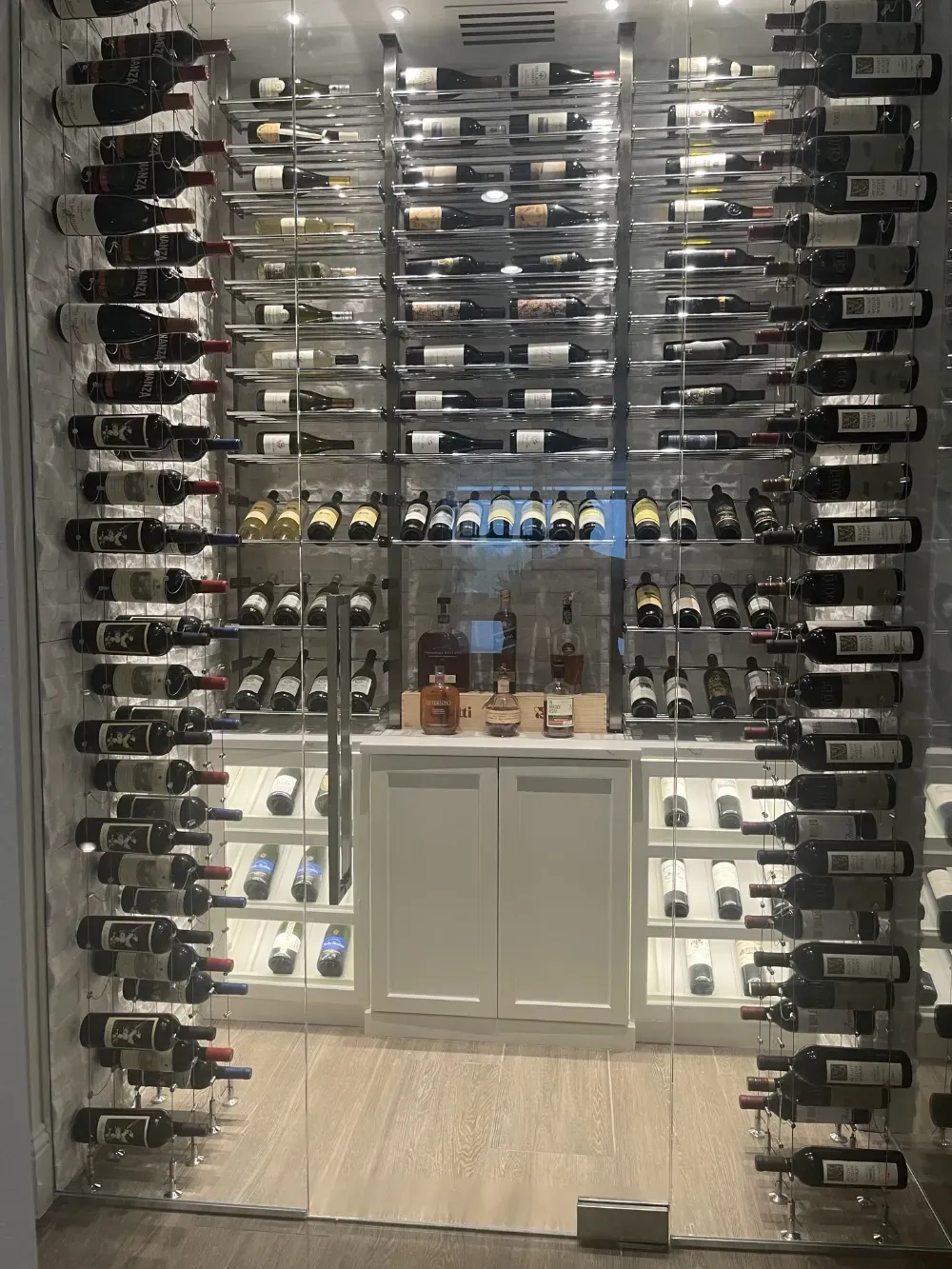 A modern, glass-enclosed wine cellar with rows of wine bottles on metal racks and a small wooden cabinet at the center.