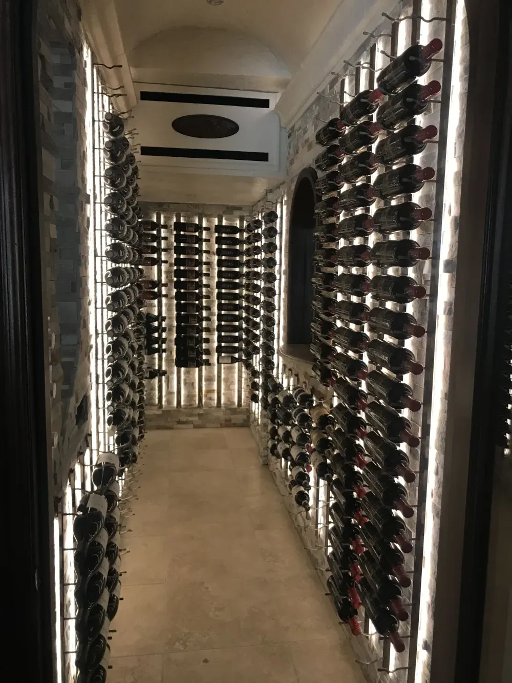 A narrow, brightly lit wine cellar featuring rows of wine bottles stored horizontally on metal wall racks.