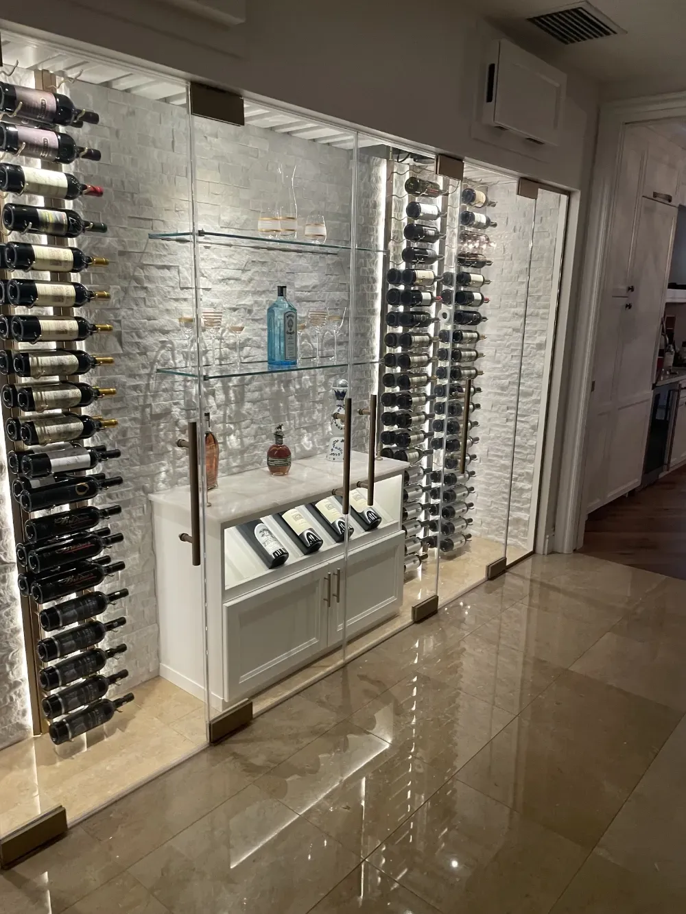 A modern, well-lit wine cellar with glass walls, horizontal bottle racks, and a central white cabinet with shelving.