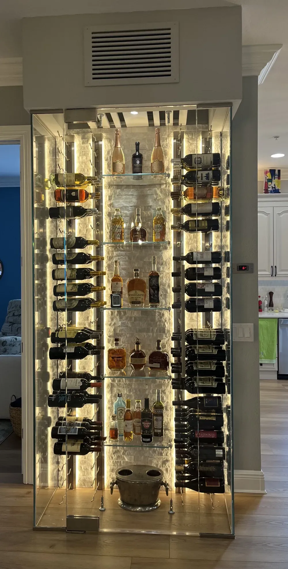 A modern, illuminated glass wine cabinet with rows of wine bottles and liquor on shelves, set in a home interior.