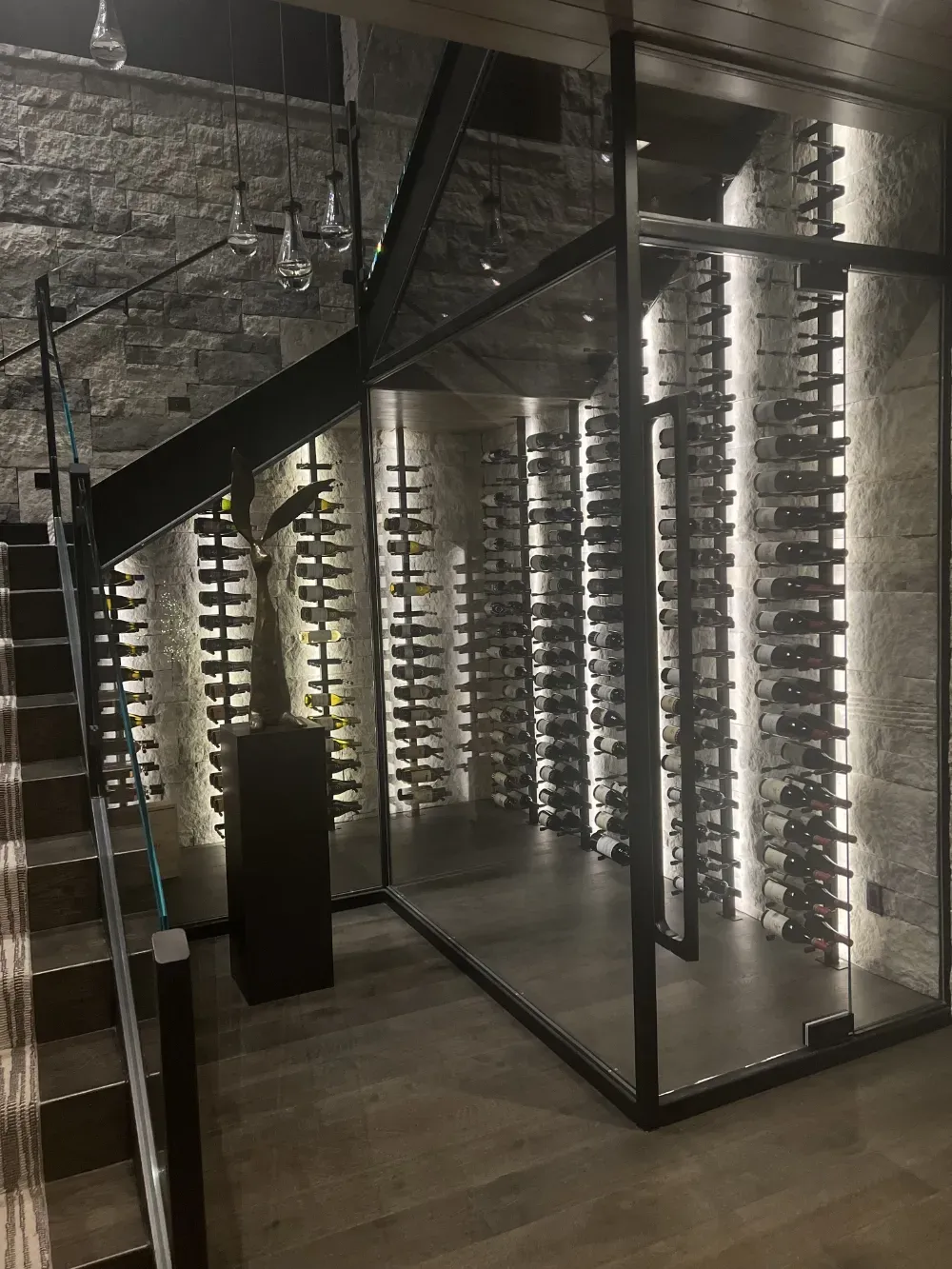 A glass-enclosed wine cellar with illuminated vertical bottle racks and a dark metal frame positioned under a staircase.