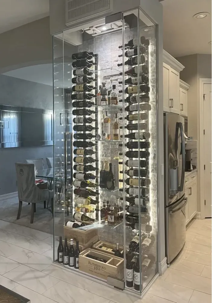 A floor-to-ceiling glass-enclosed wine display cabinet with backlit shelves, featuring organized wine bottles and decor.
