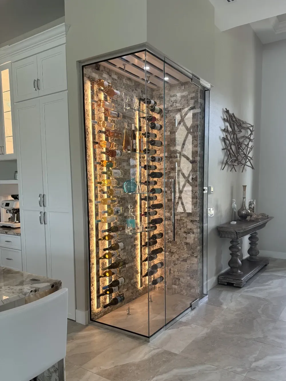 A modern, glass-enclosed wine cellar with vertical illuminated shelves, positioned in a corner of a brightly lit home.