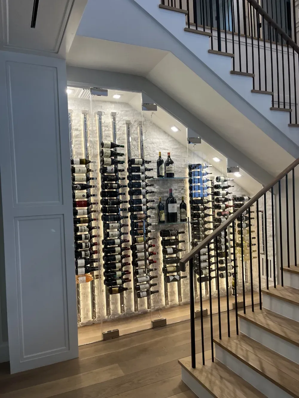 A modern, illuminated wine rack built into the storage space beneath a white wooden staircase.