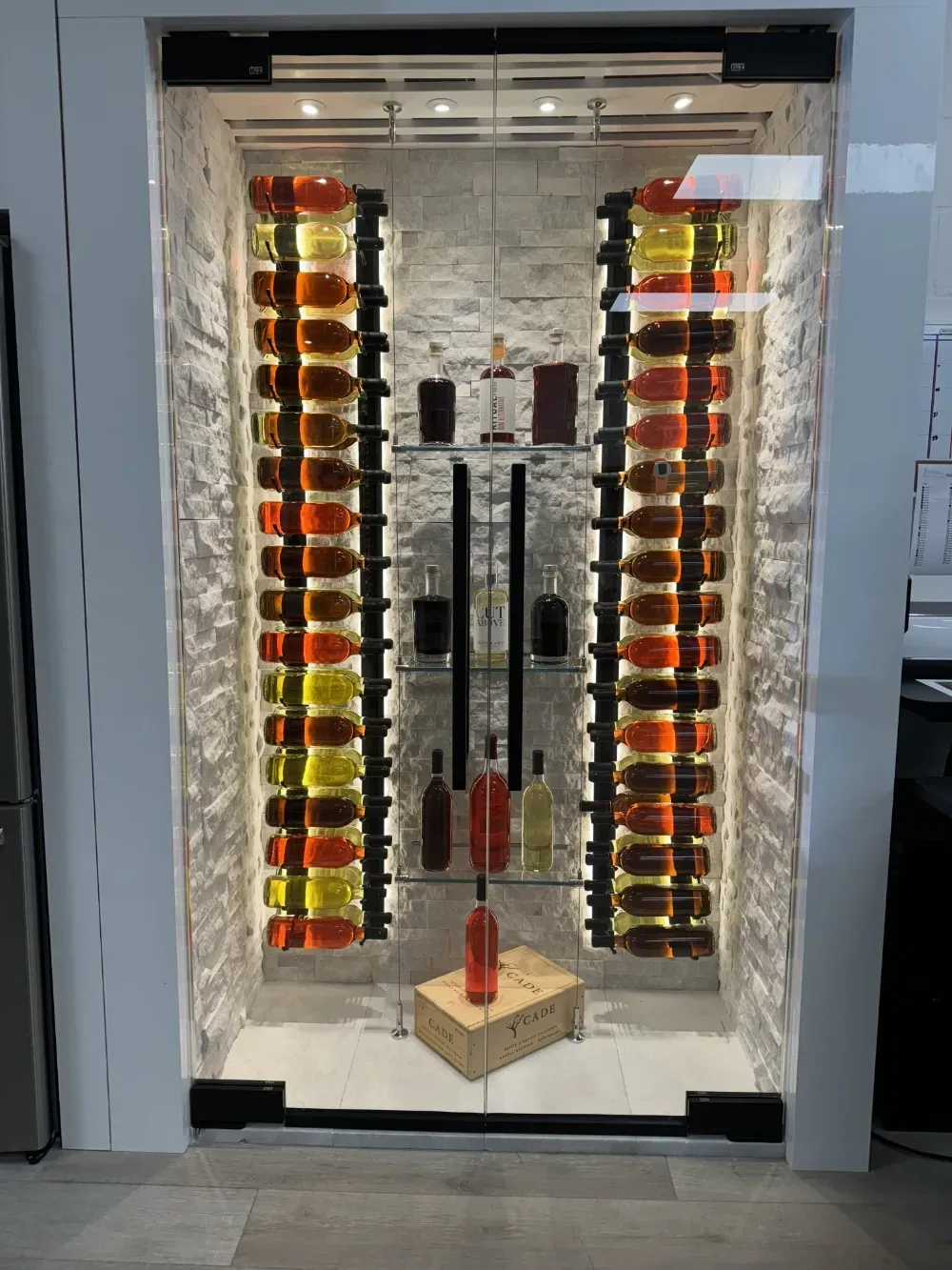 An illuminated wine display wall features two vertical racks of bottles flanking glass shelves with additional bottles.