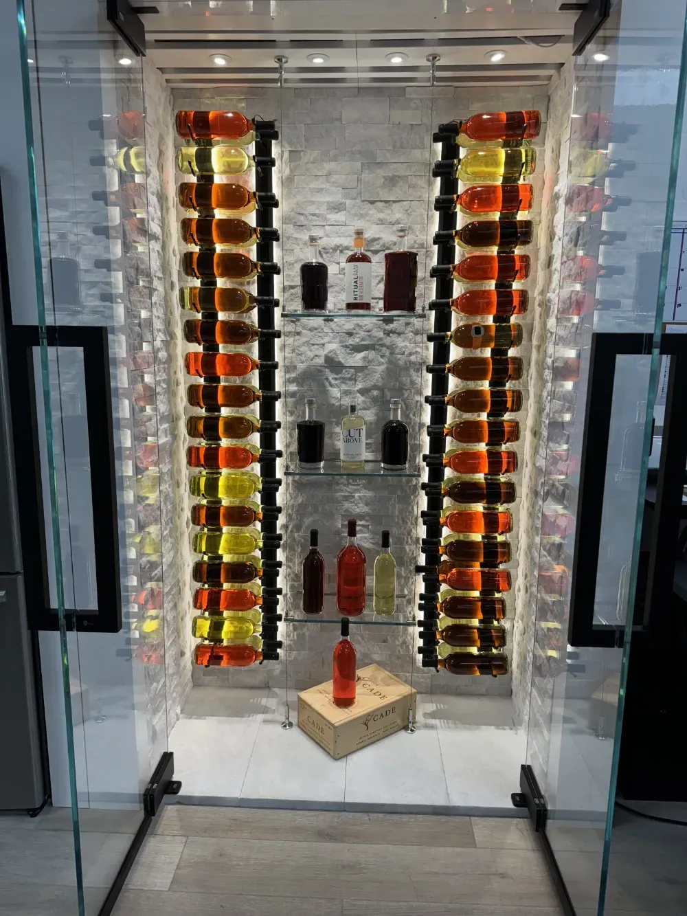 A modern glass-enclosed wine display case with side-mounted racks holding numerous bottles and central glass shelves.