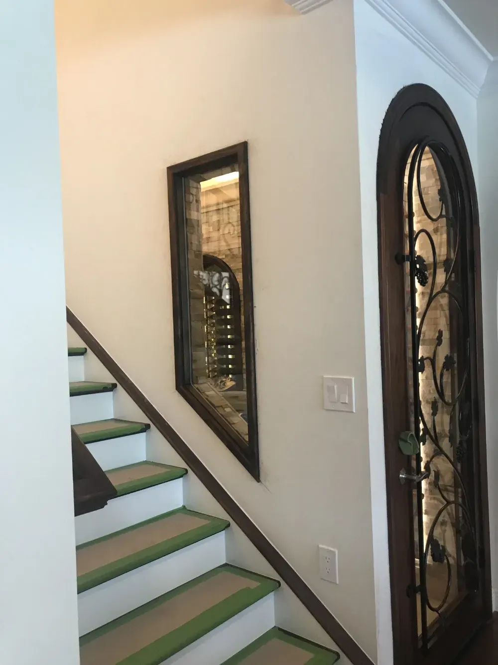 A staircase with green carpet runners beside a wall-mounted rectangular mirror and an arched door with iron scrollwork.