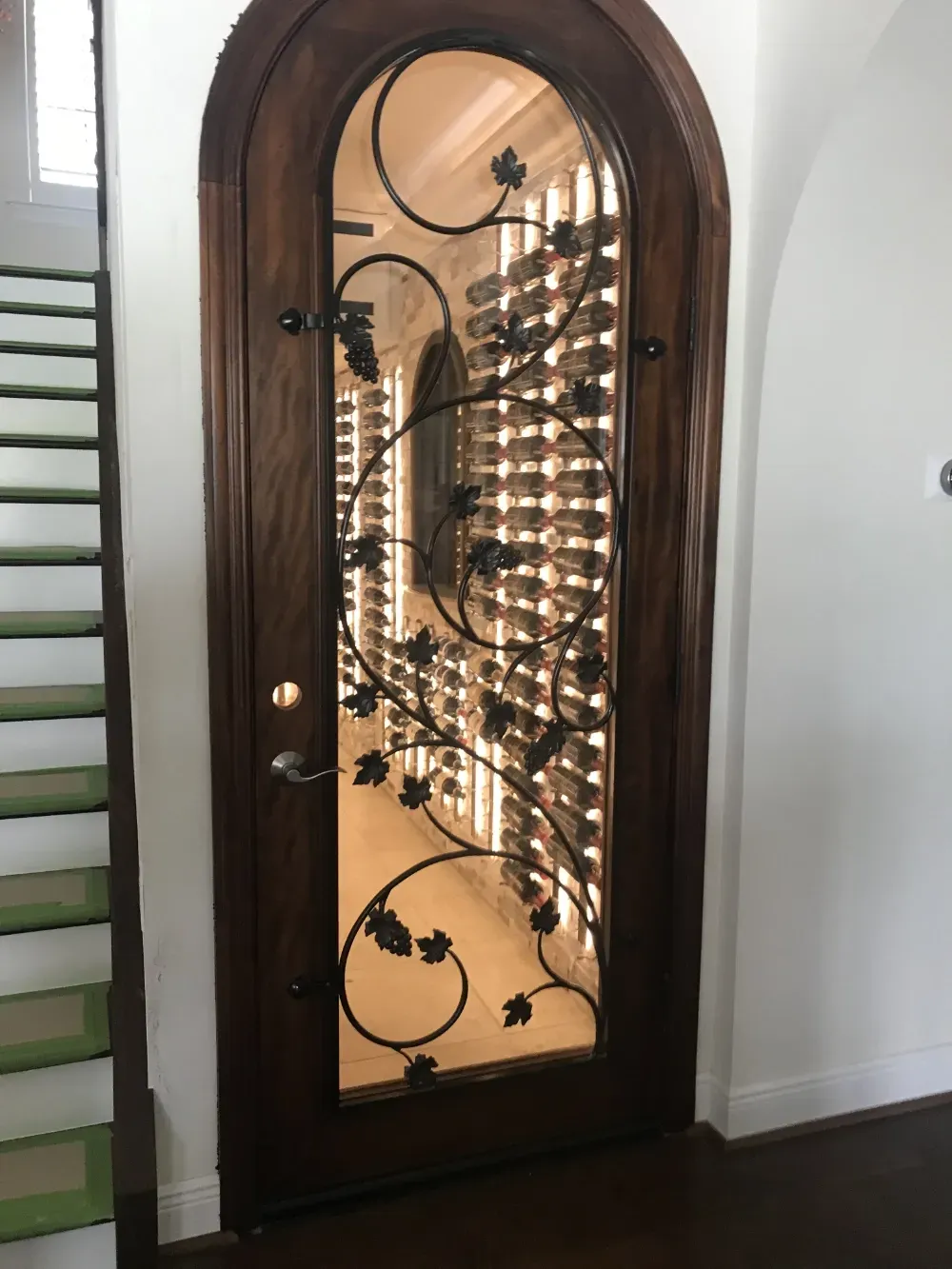 Arched dark wood door with a decorative iron vine scroll over glass, revealing a wine cellar with backlit bottle racks.