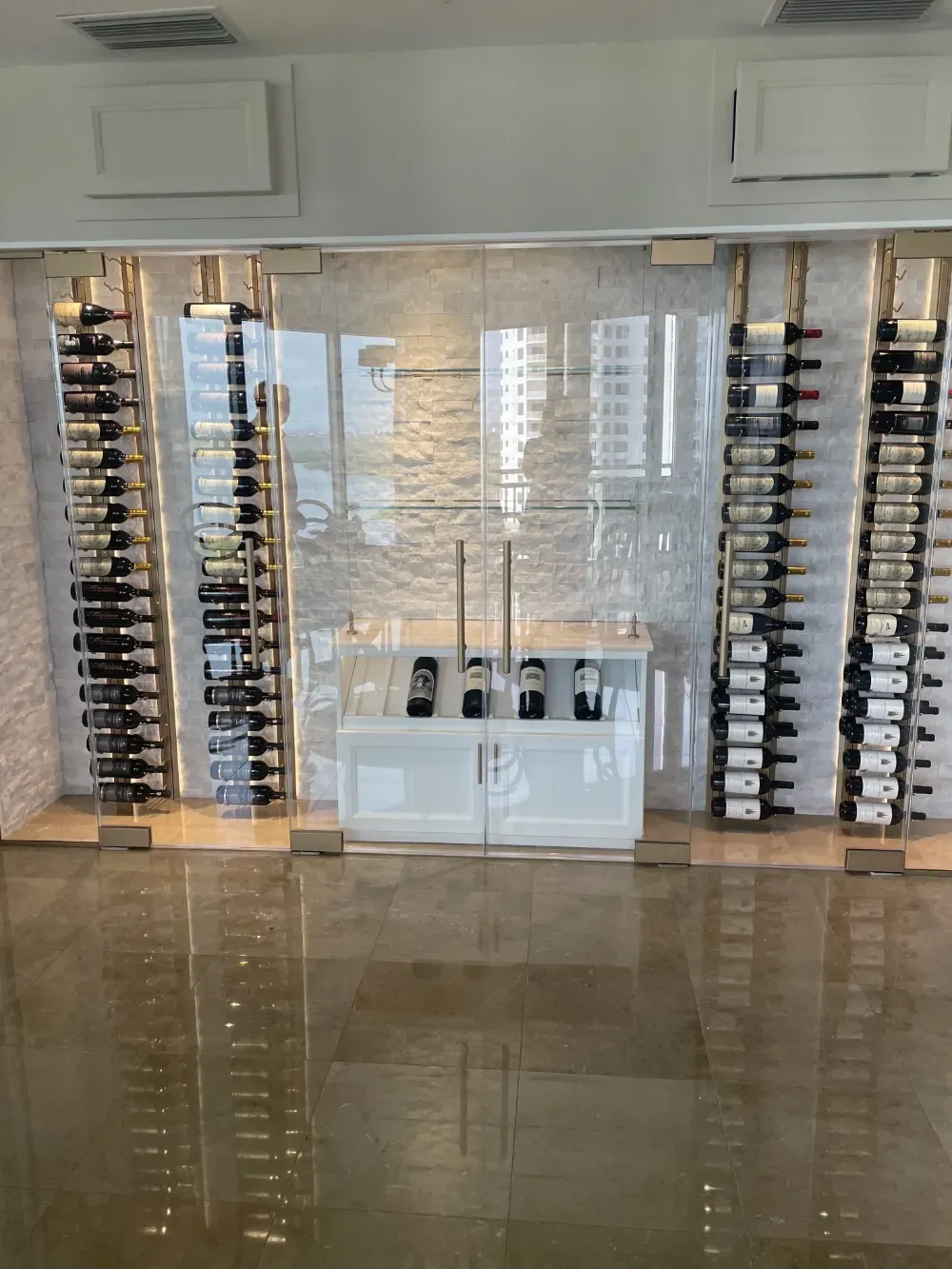 A modern, glass-enclosed wine cellar with vertical racks holding numerous wine bottles and a central white cabinet.