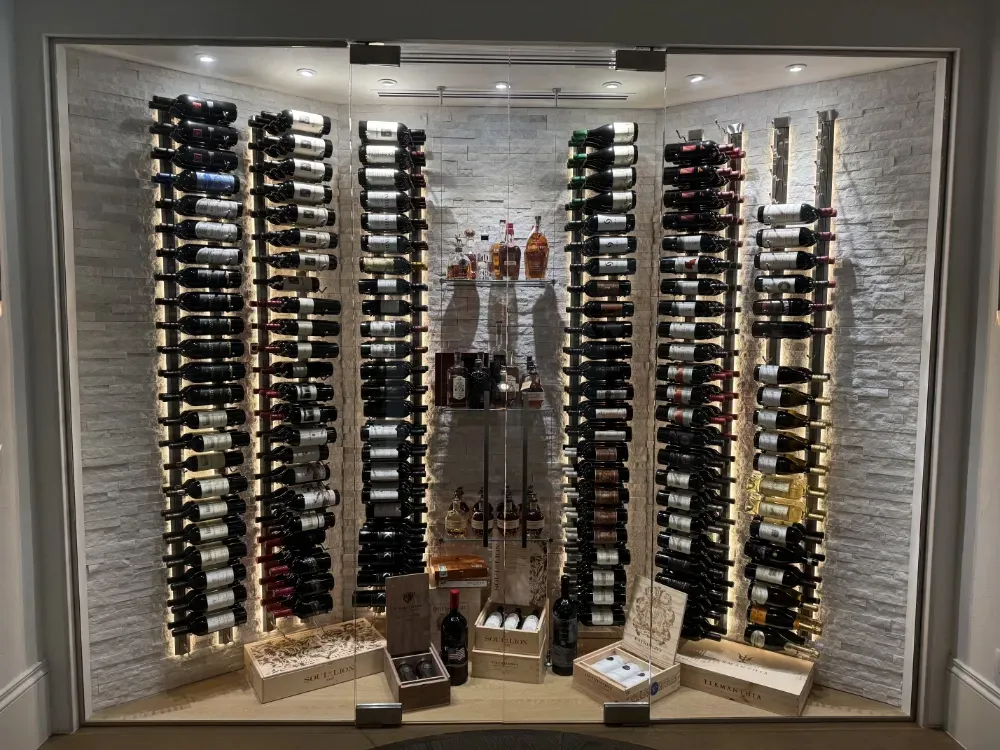 A glass-enclosed wine cellar with vertical racks holding many bottles, illuminated shelves, and crates on the floor.