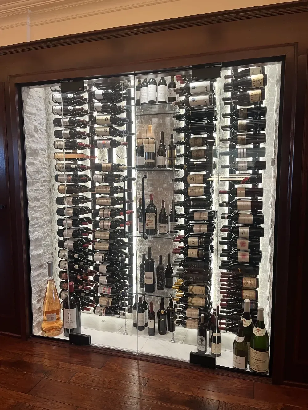 A glass-enclosed, illuminated wine cellar with multiple vertical racks showcasing a wide selection of bottled wine.