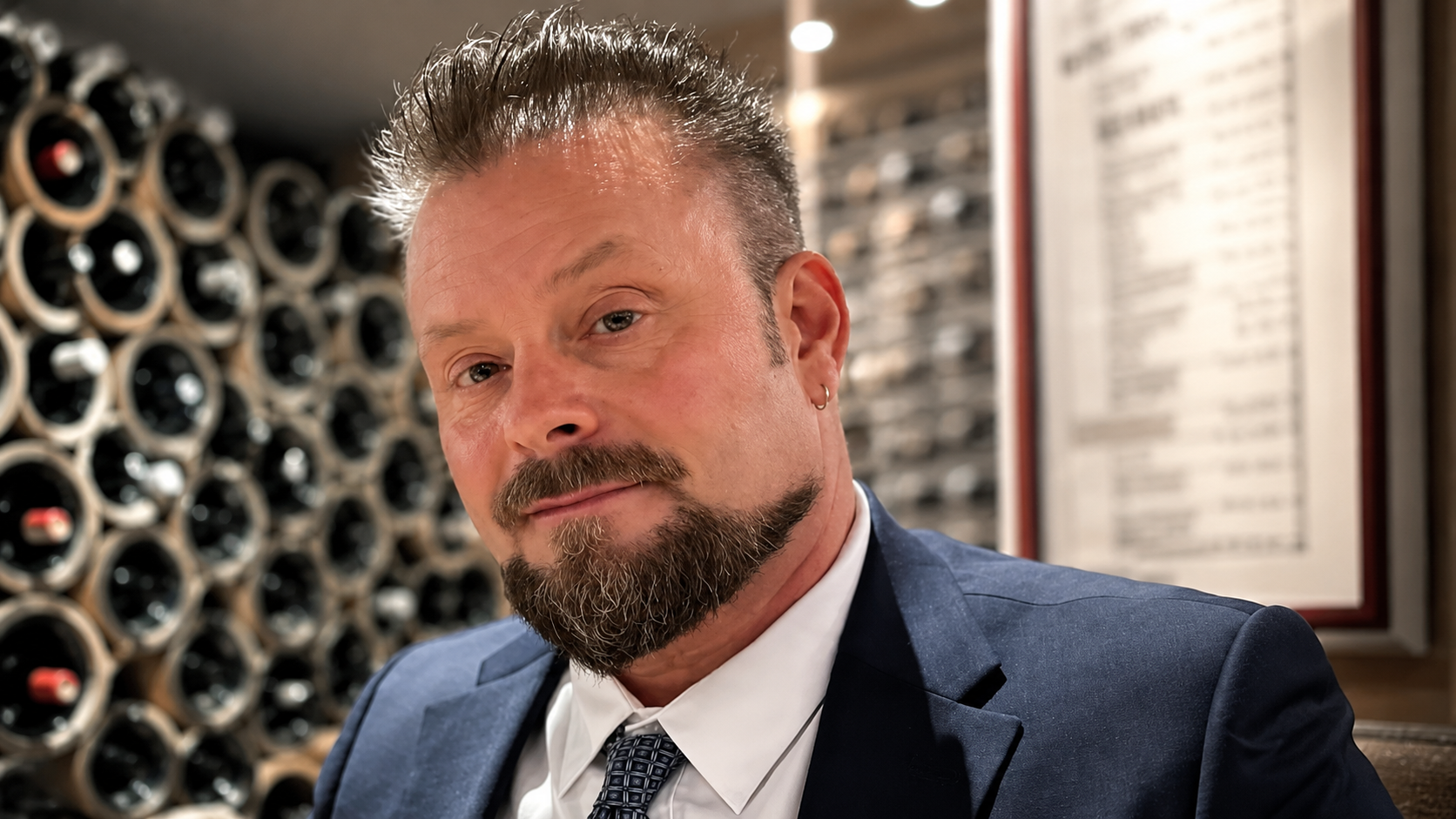 A person with a beard and spiky hair wearing a blue blazer and patterned tie, set against a wine rack background.