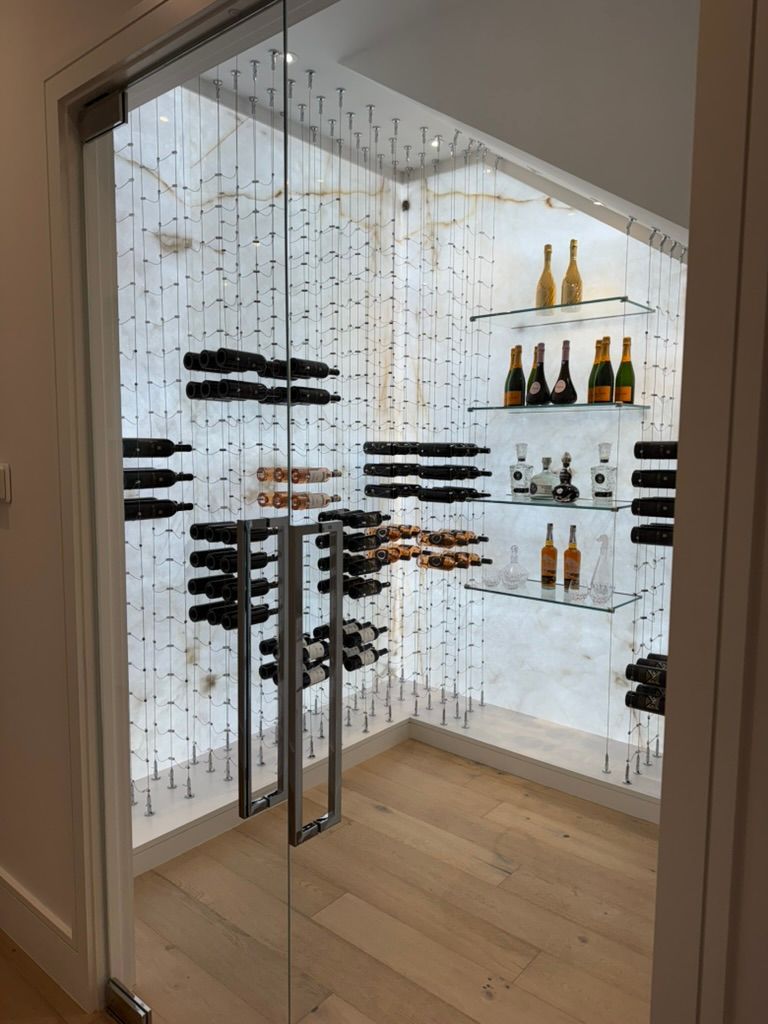 A modern, glass-enclosed wine cellar with wall-mounted bottle racks, illuminated glass shelving, and light wood flooring.