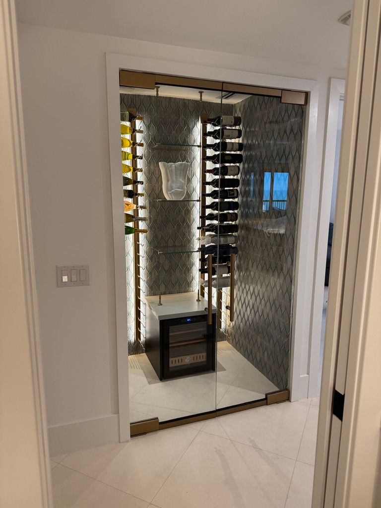 A modern, glass-enclosed wine closet with vertical racks, illuminated shelving, and a small wine fridge on white tile.
