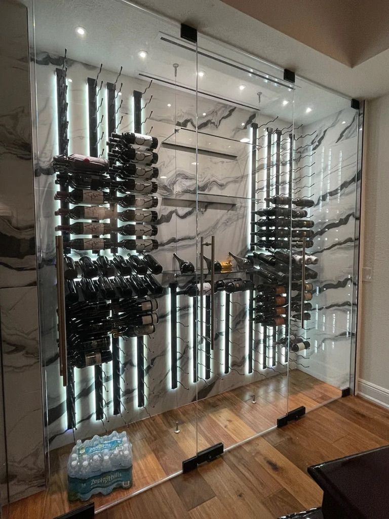 A glass-enclosed wine cellar with illuminated racks mounted on a white and gray marble wall, set on a hardwood floor.