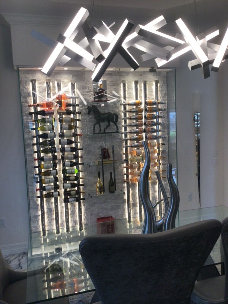 Modern wine display wall with backlit stone, metal wine racks, and an abstract geometric chandelier above a glass table.