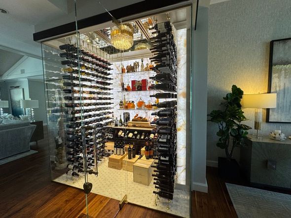 A glass-enclosed, well-lit wine room featuring extensive bottle storage racks and a central illuminated display shelf.
