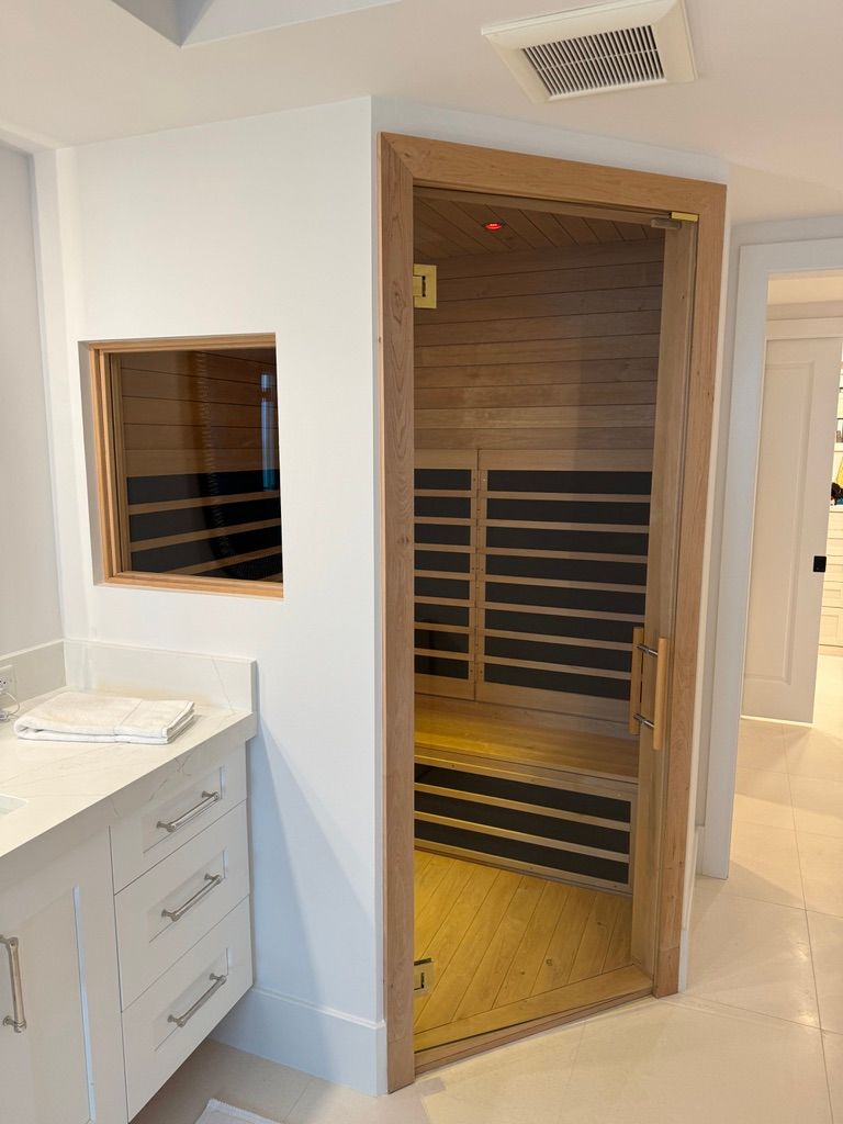 A small, modern home sauna with light wood paneling and black heating panels, built into the corner of a white bathroom.