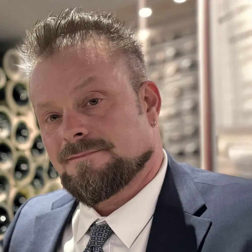 A person with a beard and spiky hair wearing a blue suit, white shirt, and patterned tie, set against a wine cellar.