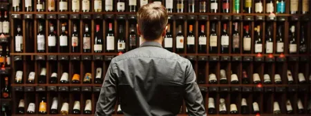A person in a gray shirt stands facing a large, well-stocked wooden wine rack in a dimly lit cellar.