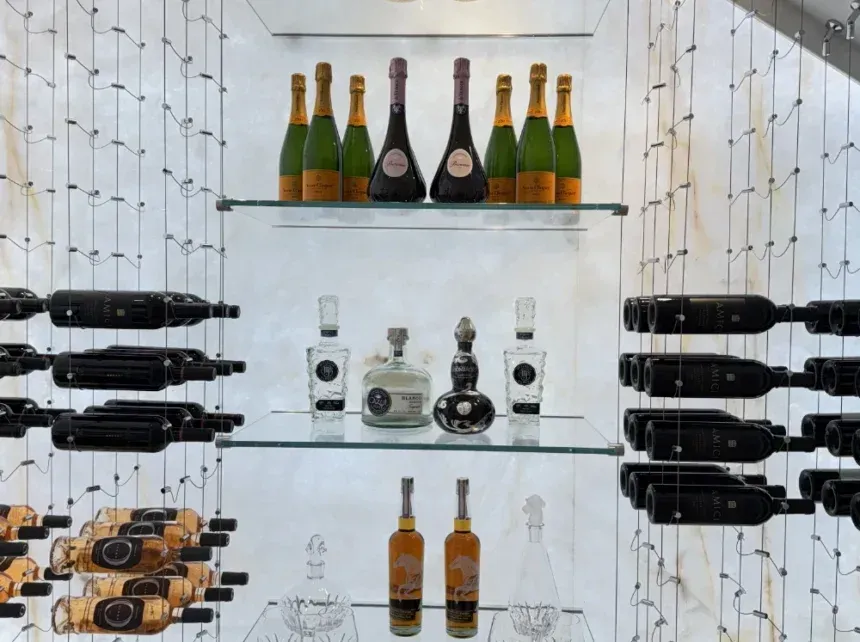 A wine cellar display with glass shelves holding spirits and champagne, flanked by bottles stored on hanging metal racks.