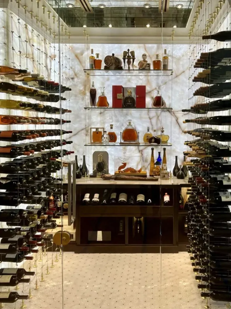 A modern, brightly lit wine cellar with glass-enclosed shelves showcasing wine bottles and decorative liquor decanters.