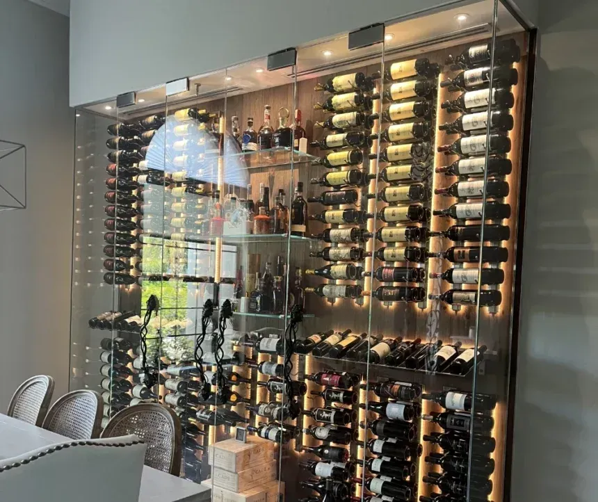A modern, floor-to-ceiling glass wine cellar cabinet with interior LED lighting, displaying rows of bottles and liquor.
