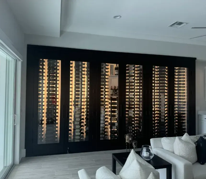 A wall-to-wall, black-framed wine cellar with illuminated vertical display racks, set in a modern, light-toned living room.