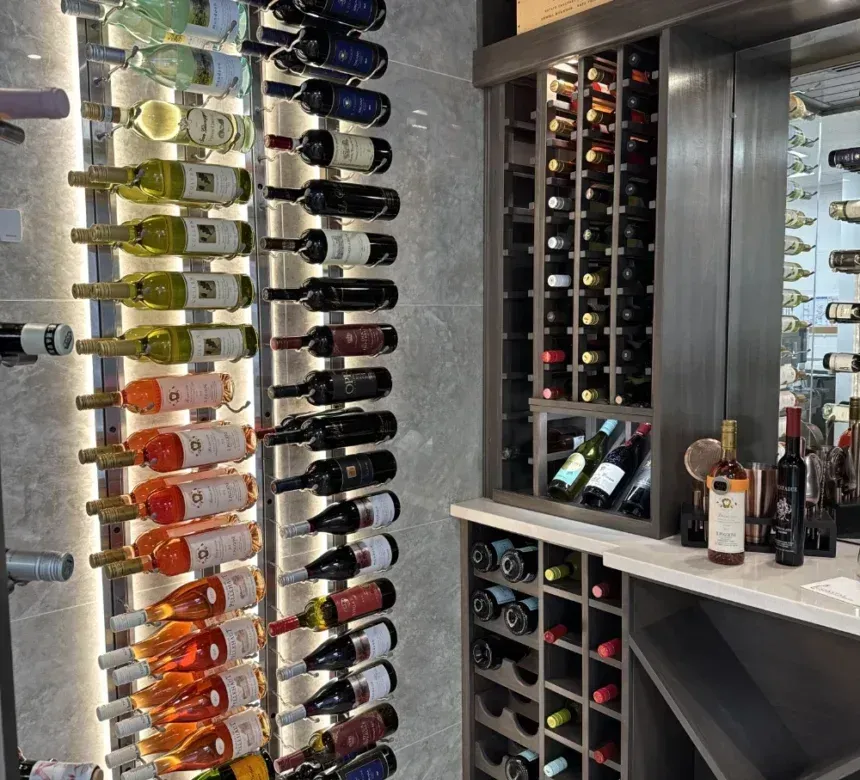 A wine cellar wall with illuminated vertical racks holding many bottles, next to a wooden grid rack and a serving counter.