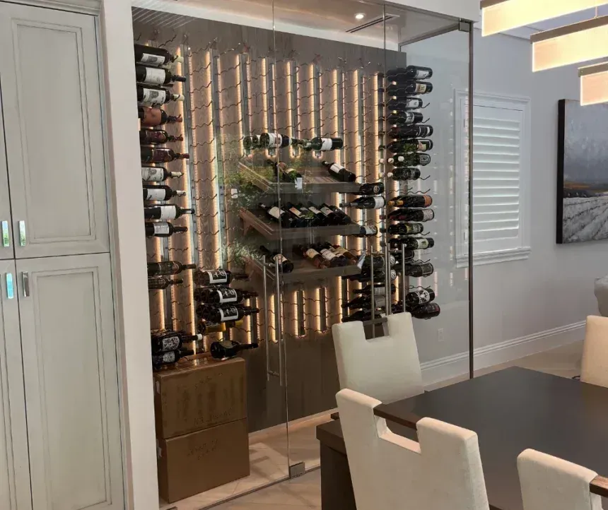A glass-enclosed wine display unit with metal racks mounted against a vertically lit wood-paneled wall, near a dining set.