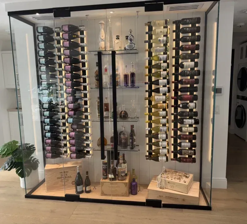 A modern, illuminated glass wine display unit filled with rows of wine bottles and liquor on central shelves.