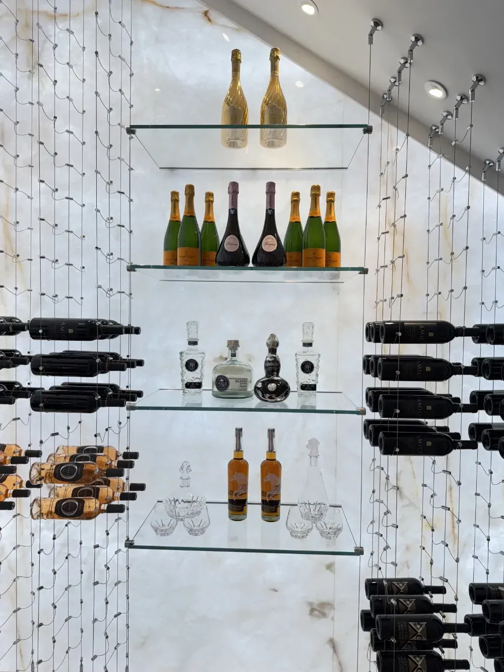 Wine bottles on glass shelves in a wine cellar. Champagne, liquors, and red wine are displayed.