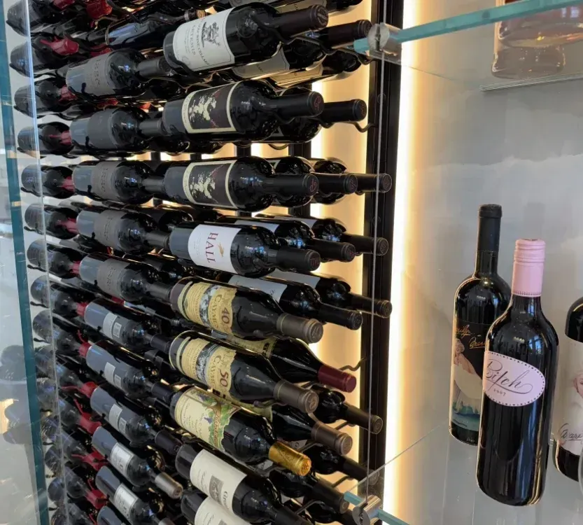 Wine bottles stored horizontally on metal racks inside a glass-enclosed display case, with some bottles on a side shelf.